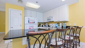 A kitchen with white cabinets, a refrigerator, a microwave, and a dining area with a bar stool and chairs. - LIGHTHOUSE! Popular Property! ~ 1 BR Beach Front! XTRA BATH! (Gulf Shores)