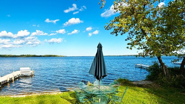 A lakeside outdoor seating area with a round table and chairs, an umbrella, and a dock extending into the water. - Lakeside Cabin on Lake Noquebay - Including Your Own Pier, Kayak/Canoes, & More! (Crivitz)