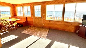 A cabin with a wooden interior, a dining area with a table and chairs, a television, and large windows offering a view of the water. - Lakeside Cabin on Lake Noquebay - Including Your Own Pier, Kayak/Canoes, & More! (Crivitz)