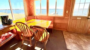 A wooden cabin with a dining table and chairs, large windows overlooking a body of water, and a check-out sign on the door. - Lakeside Cabin on Lake Noquebay - Including Your Own Pier, Kayak/Canoes, & More! (Crivitz)