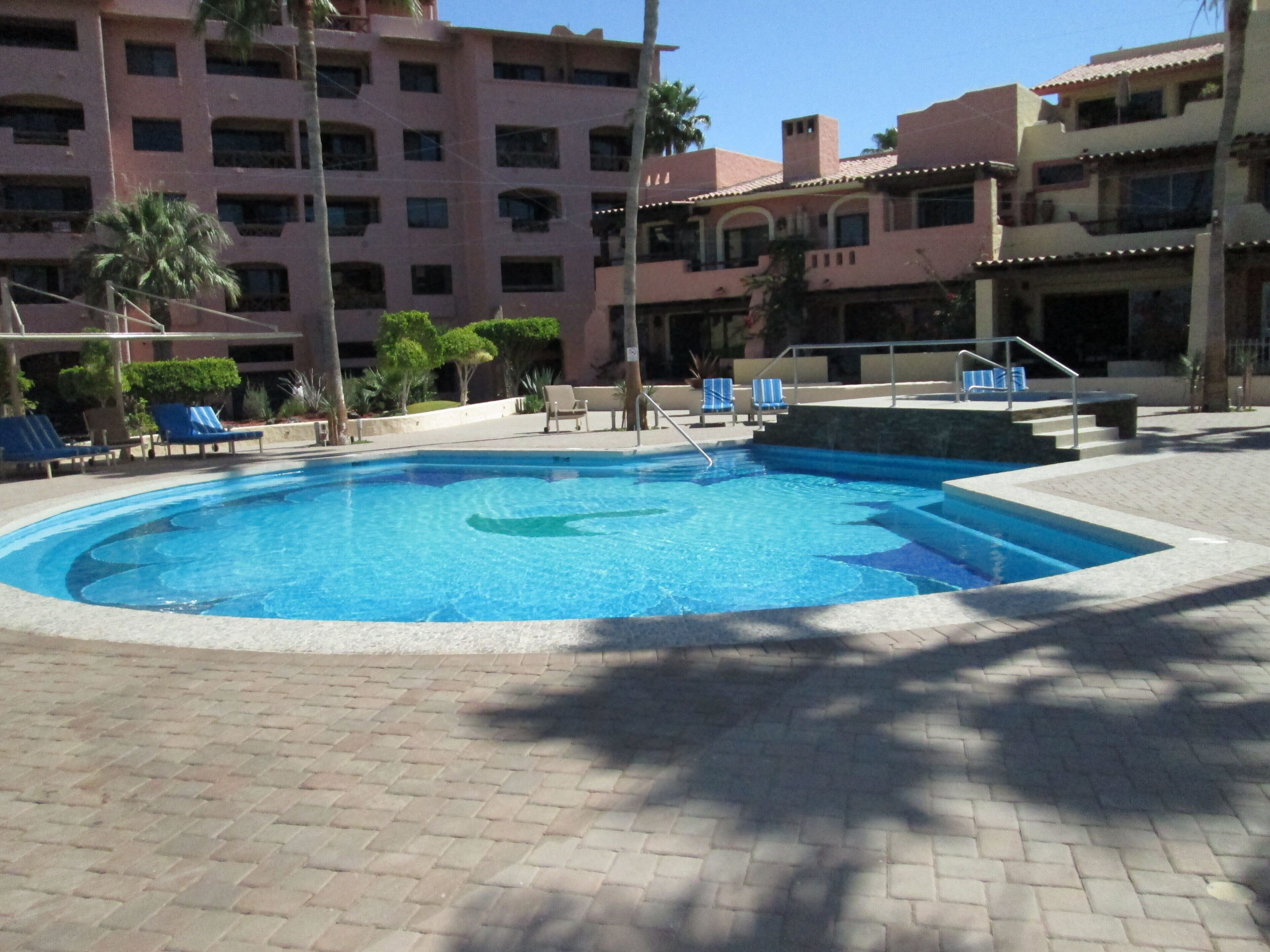 A swimming pool with lounge chairs, a stone pathway, and a multi-story building in the background.