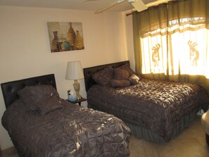 A hotel room with two beds, a bedside table with a lamp, a painting on the wall, and a window with curtains.