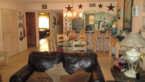 A living room with a black leather sofa, a dining table with chairs, a chandelier, and a kitchen area in the background.