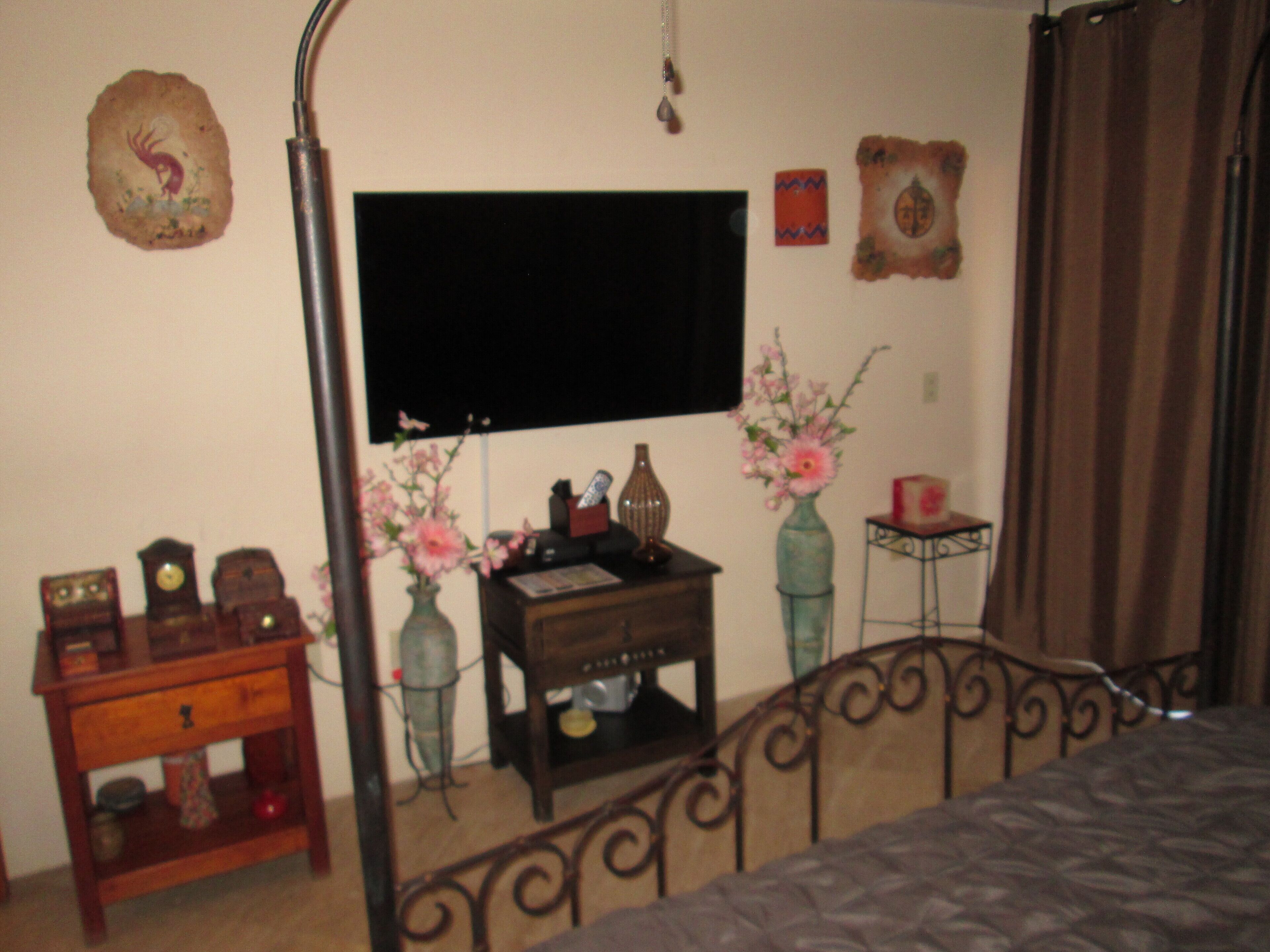 Ein Schlafzimmer mit Bett, einem an der Wand montierten Fernseher, einem kleinen Holztisch mit Blumenvase und einer dekorativen Wandgestaltung.