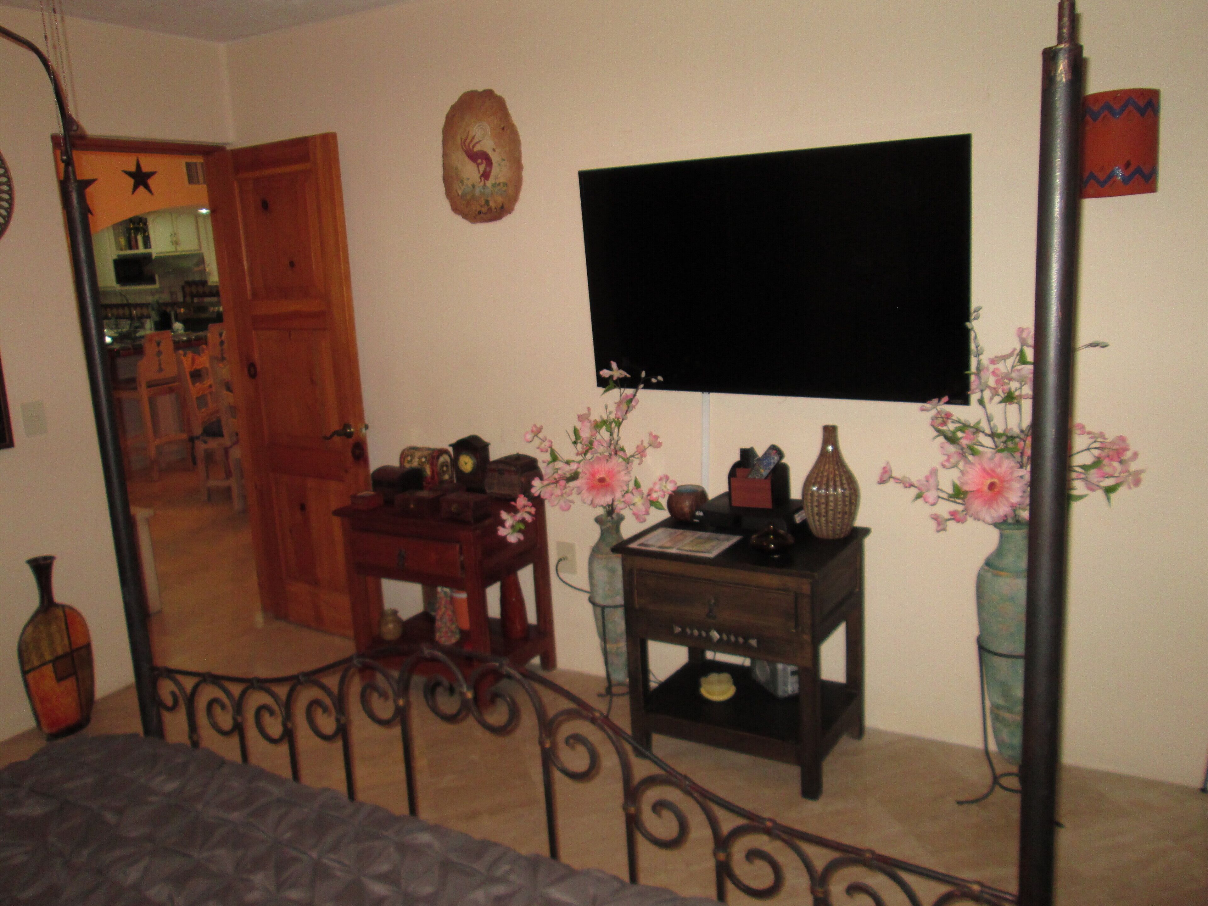Una sala de estar con un televisor montado en la pared, una mesita de madera con varios objetos, un florero con flores y una puerta que da a otra habitación.