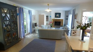 Ein Wohnzimmer mit einer Couch, einem Esstisch und einem Fernseher. Es gibt ein Fenster mit Vorhängen, eine Pflanze und ein Gemälde an der Wand.