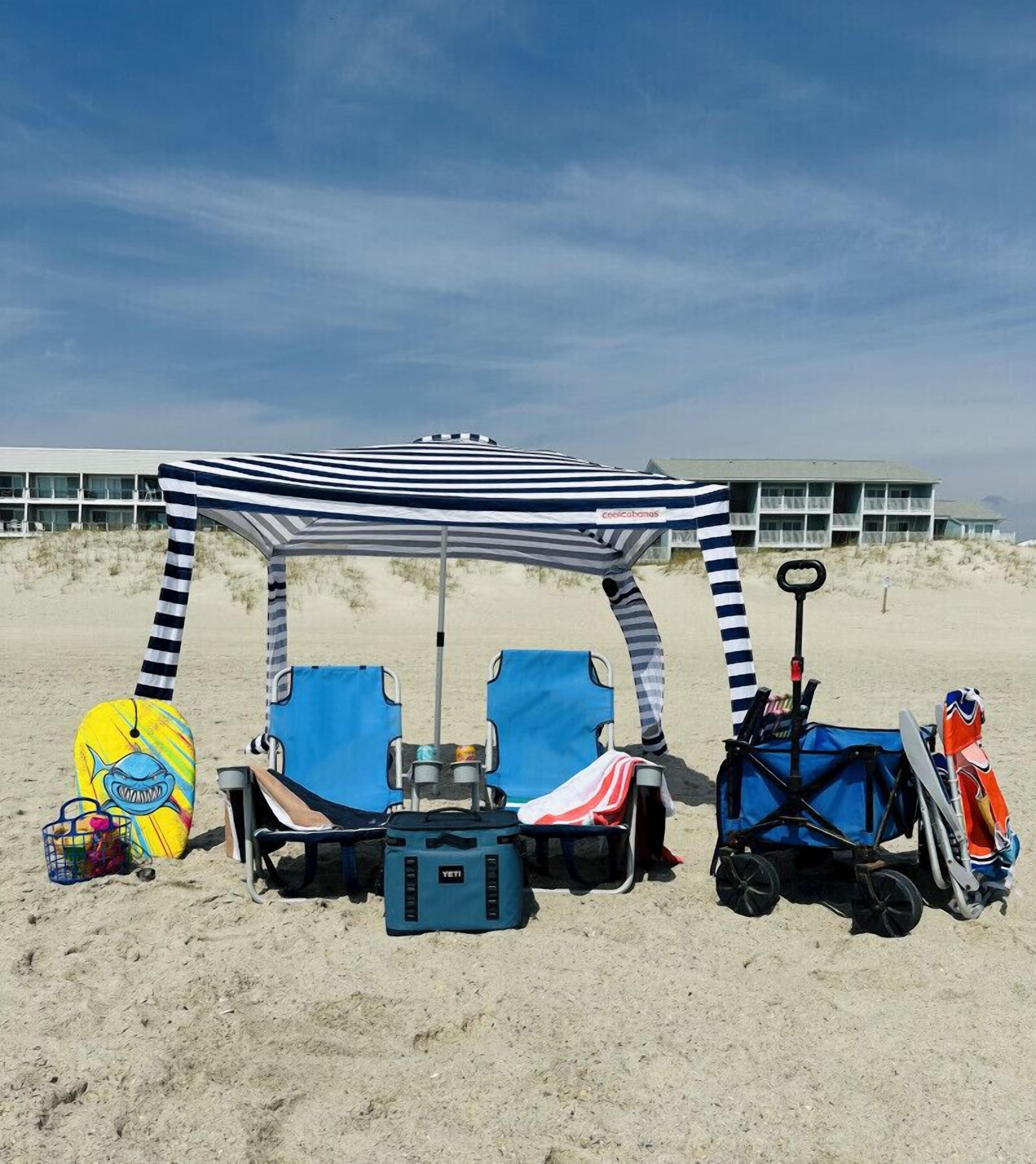 A beach setup with a striped canopy, two blue beach chairs, a blue cooler, a beach bag, and a stroller.
