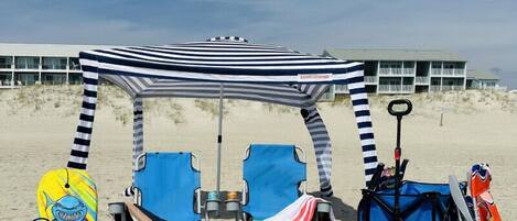 A beach setup with a striped canopy, two blue beach chairs, a blue cooler, a beach bag, and a stroller.