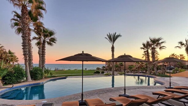 A poolside area with lounge chairs and umbrellas, overlooking a body of water and palm trees.