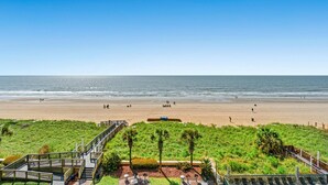 Una playa con un paseo marítimo de madera que conduce a una zona verde con bancos y una pequeña construcción.