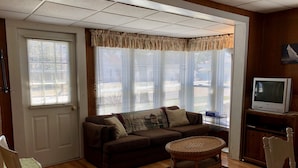 A living room with a brown sofa, a round wicker table, a television on a wooden stand, and a door with a window.