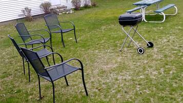 Une cour arrière verdoyante avec un barbecue, une table de pique-nique et des chaises d’extérieur.