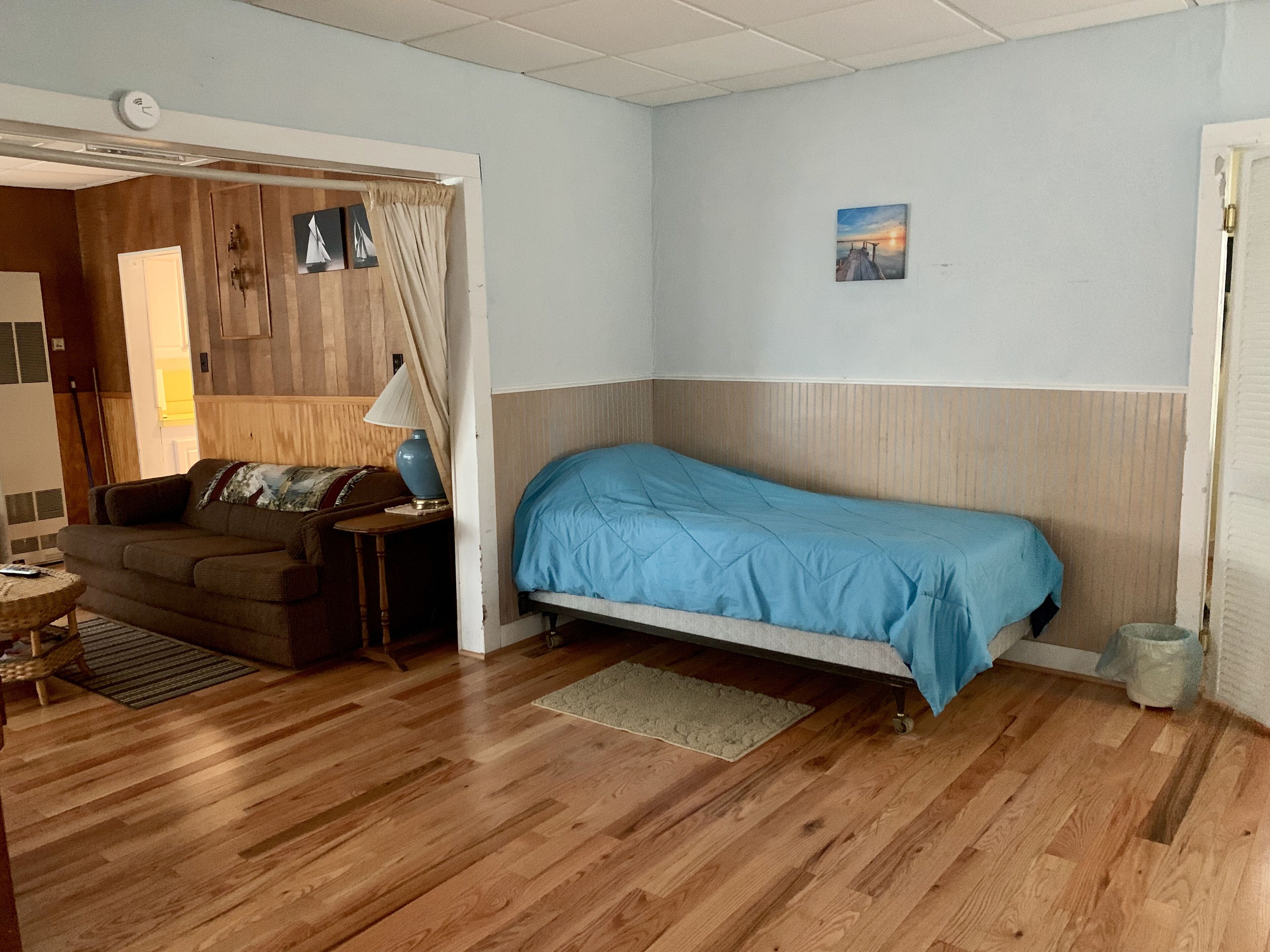 A bedroom with a single bed, a wooden floor, a small table with a lamp, a sofa, and a painting on the wall.