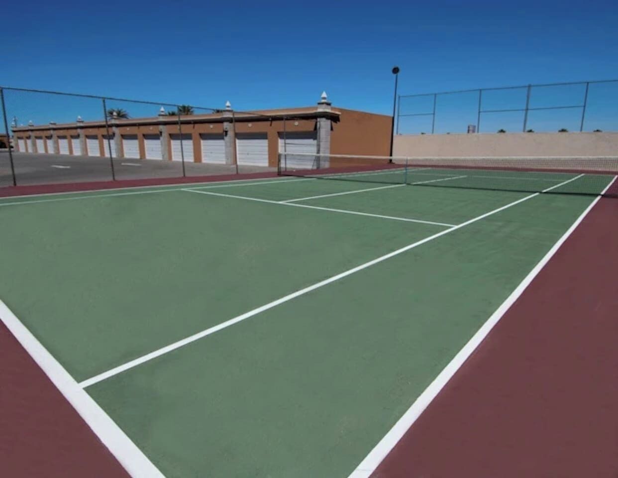Una cancha de tenis con superficie verde y bordes rojos, rodeada por una valla y un edificio con múltiples puertas de garaje al fondo.