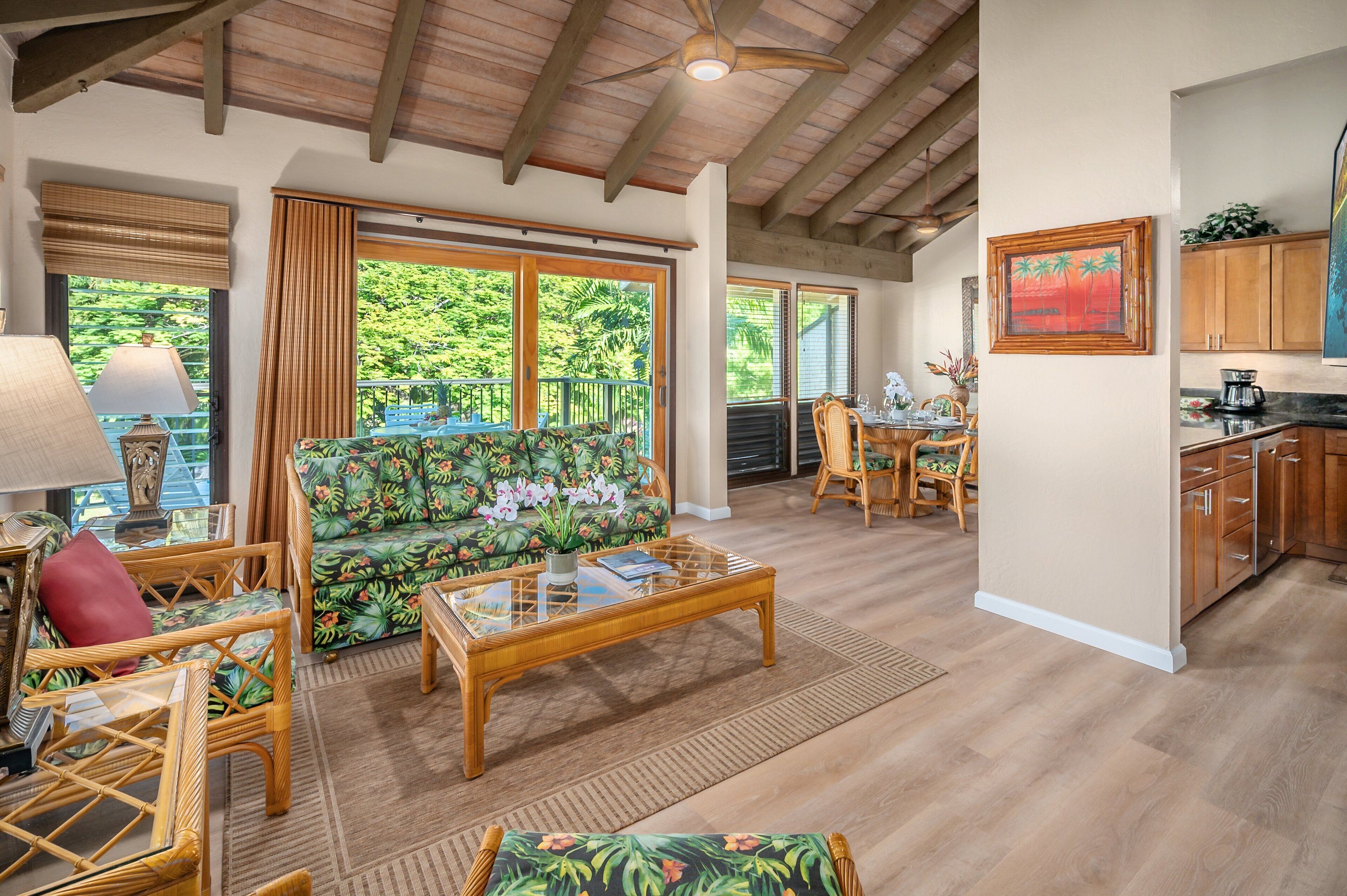 A spacious living area with a sofa, coffee table, and wicker chairs. A dining area with a table and chairs is visible in the background. The room has large windows with curtains, wooden floors, and a ceiling fan.