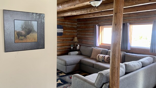 Interior de una cabaña de troncos con un sofá seccional gris, techo de madera y un cuadro enmarcado de un alce en la pared.