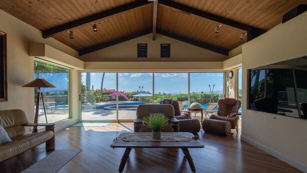 A spacious living room with a high ceiling, large windows offering a view of a pool and garden, and a cozy seating area with a coffee table. - Breath taking ocean and volcano views! Pool,Close to beaches and everything else (Kihei)