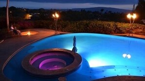 A swimming pool at dusk with a circular hot tub in the center, surrounded by a landscaped area with trees and a view of distant hills. - Breath taking ocean and volcano views! Pool,Close to beaches and everything else (Kihei)