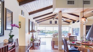 A spacious dining area with a high ceiling and wooden beams, a large window offering a view of the outdoors, and a dining table set for a meal. - Breath taking ocean and volcano views! Pool,Close to beaches and everything else (Kihei)