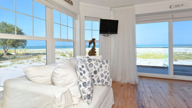 A room with a white couch, patterned pillows, a mounted TV, and large windows with a beach view. -  Beautiful, Gulf-Front Home on Bean Point--Halcyon House (Anna Maria)
