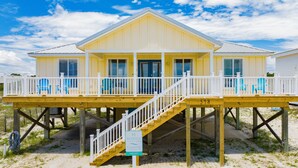 Ein zweistöckiges Strandhaus mit einer Holzterrasse, blauen Stühlen und einer Treppe.