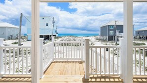 Eine Holzterrasse mit weißen Geländern, Blick auf den Strand und Gebäude im Hintergrund.