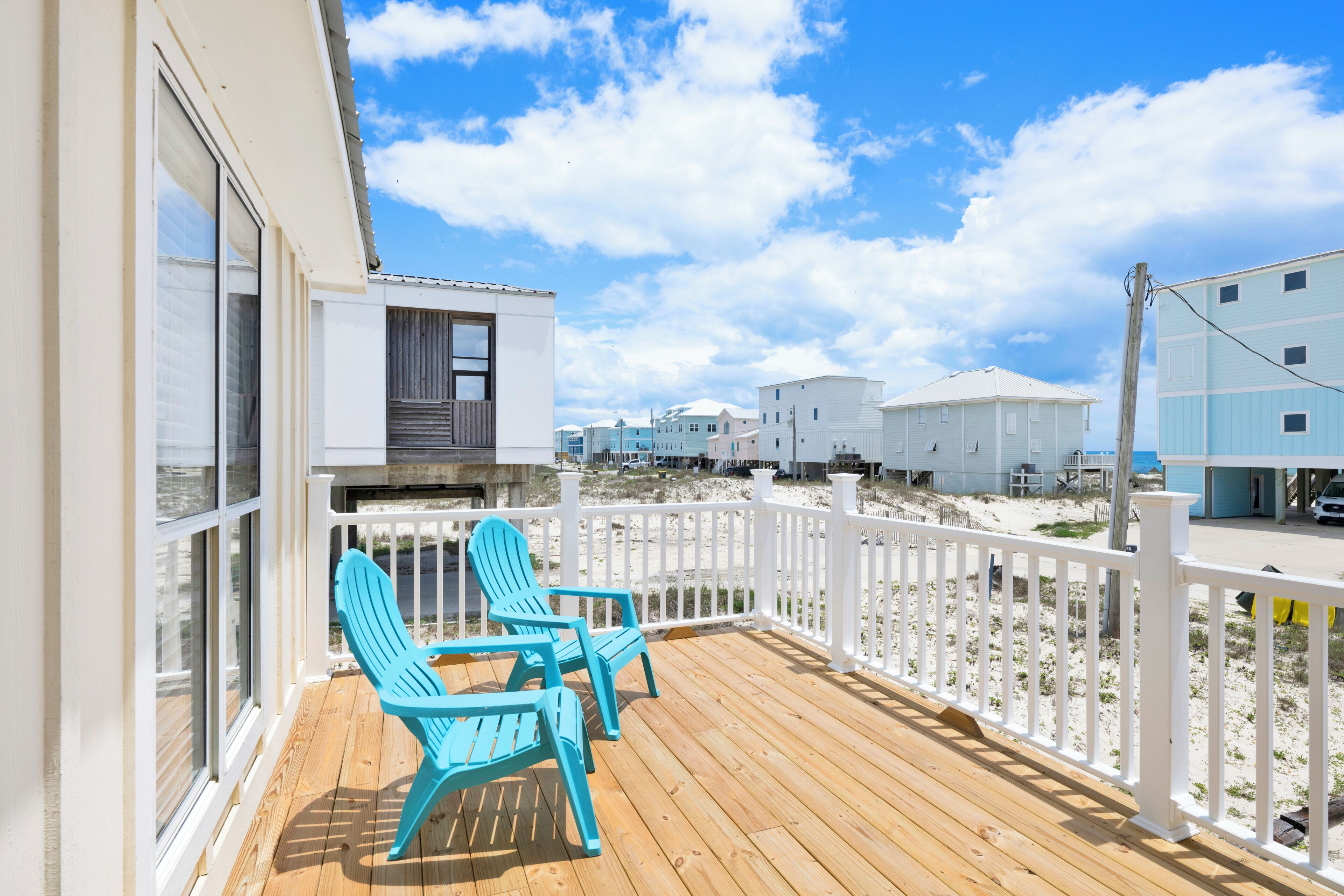 Une terrasse en bois avec deux chaises Adirondack bleues, une clôture blanche et des maisons de plage en arrière-plan.