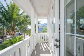 Un porche cubierto con barandillas blancas, vistas a palmeras y un ventanal de vidrio que da a una habitación.
