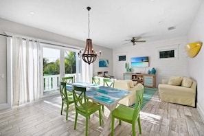 Un vaste espace de vie comprenant une table à manger, des chaises vertes, un canapé beige, un téléviseur et un balcon agrémenté de rideaux.