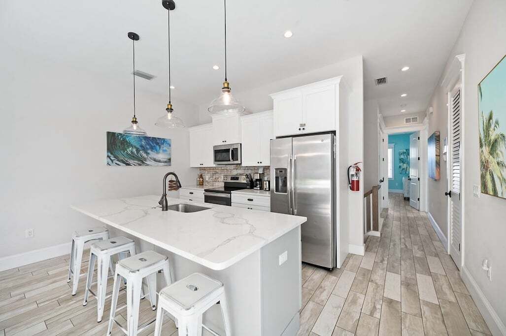 A modern kitchen with a large island, stainless steel appliances, pendant lighting, and a view into the adjacent living area.