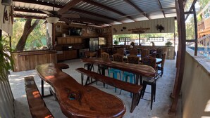 A rustic outdoor dining area with wooden benches and tables, a kitchen with wooden cabinets, and a view of a swimming pool.