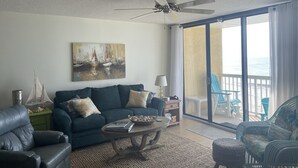 Ein Wohnzimmer mit Sofa, Couchtisch und einem Gemälde von Booten. Es gibt einen Deckenventilator, Meerblick durch große Fenster und Gartenmöbel.