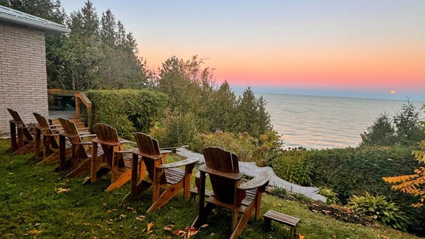 A row of wooden Adirondack chairs facing a body of water with a sunset in the background. - Peach of a private beach! Waterfront cliff cottage near Bayfield (Zurich)
