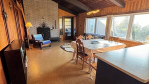 A cozy interior with a wooden ceiling, a brick fireplace, a dining area with a table and chairs, and a view through large windows. - Peach of a private beach! Waterfront cliff cottage near Bayfield (Zurich)