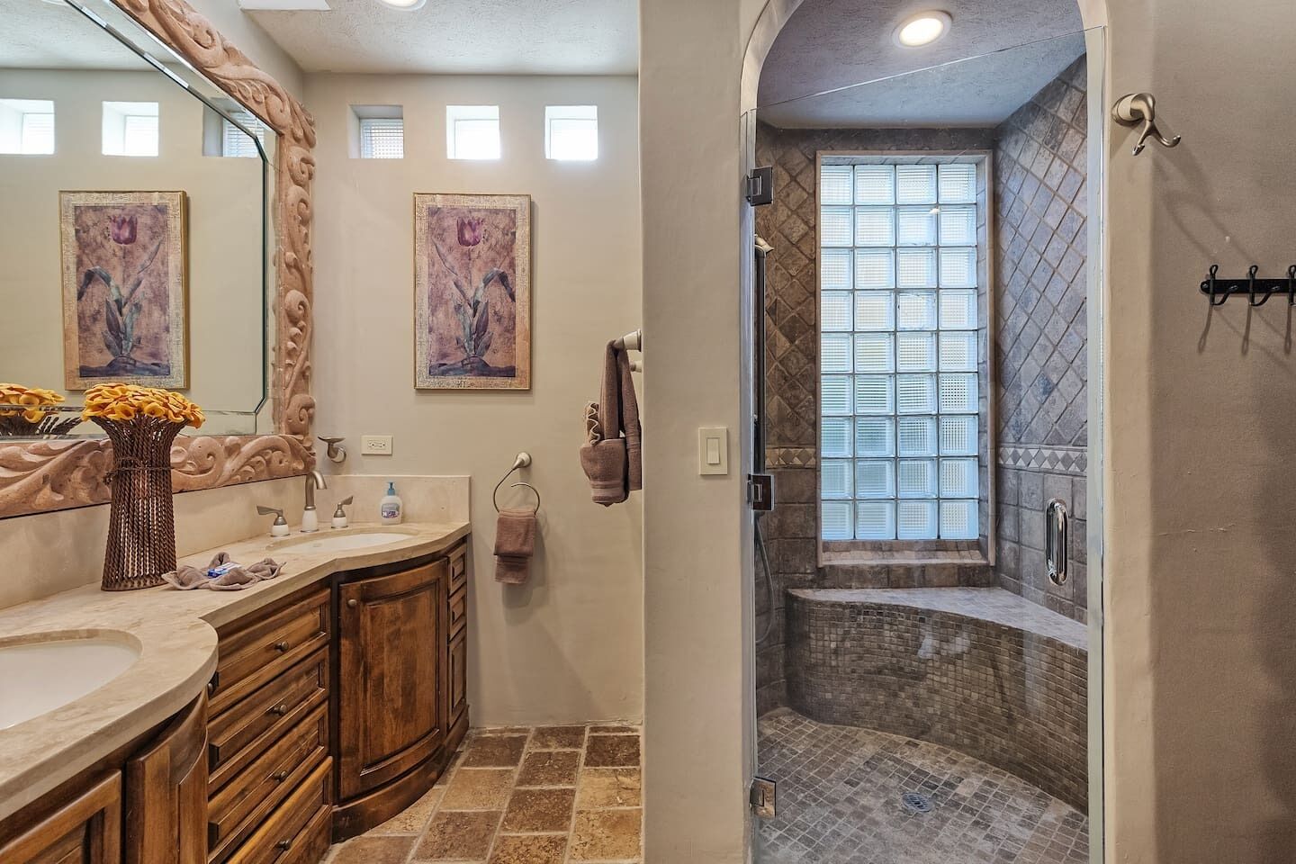 A bathroom with a vanity, a large mirror, two framed paintings, a vase with flowers, and a shower with a tiled wall and window.
