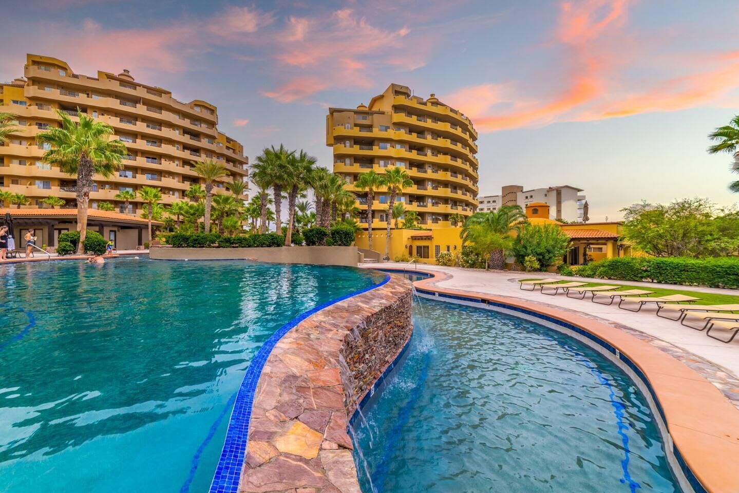 A swimming pool with a stone edge, surrounded by palm trees and a hotel with multiple balconies.