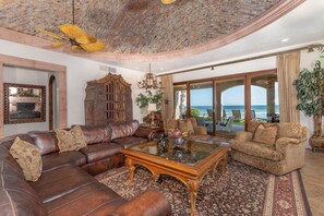 Uma sala de estar ampla com sofá de couro, mesa de centro e vista para o oceano através de grandes portas deslizantes.
