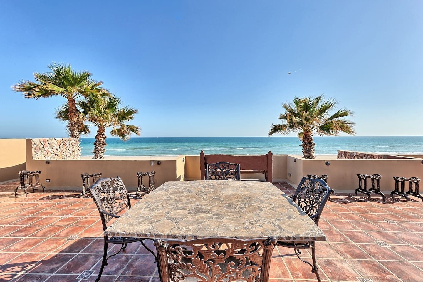 A terrace with a stone table and chairs, palm trees, and a view of the sea.