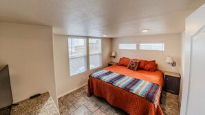 A bedroom with a large bed, two windows with blinds, a nightstand, and a lamp.