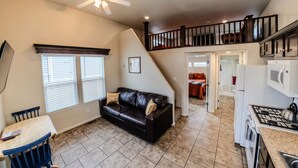 A compact living space with a black leather sofa, a dining table with chairs, a kitchen area with a stove and microwave, and a staircase leading to an upper level.
