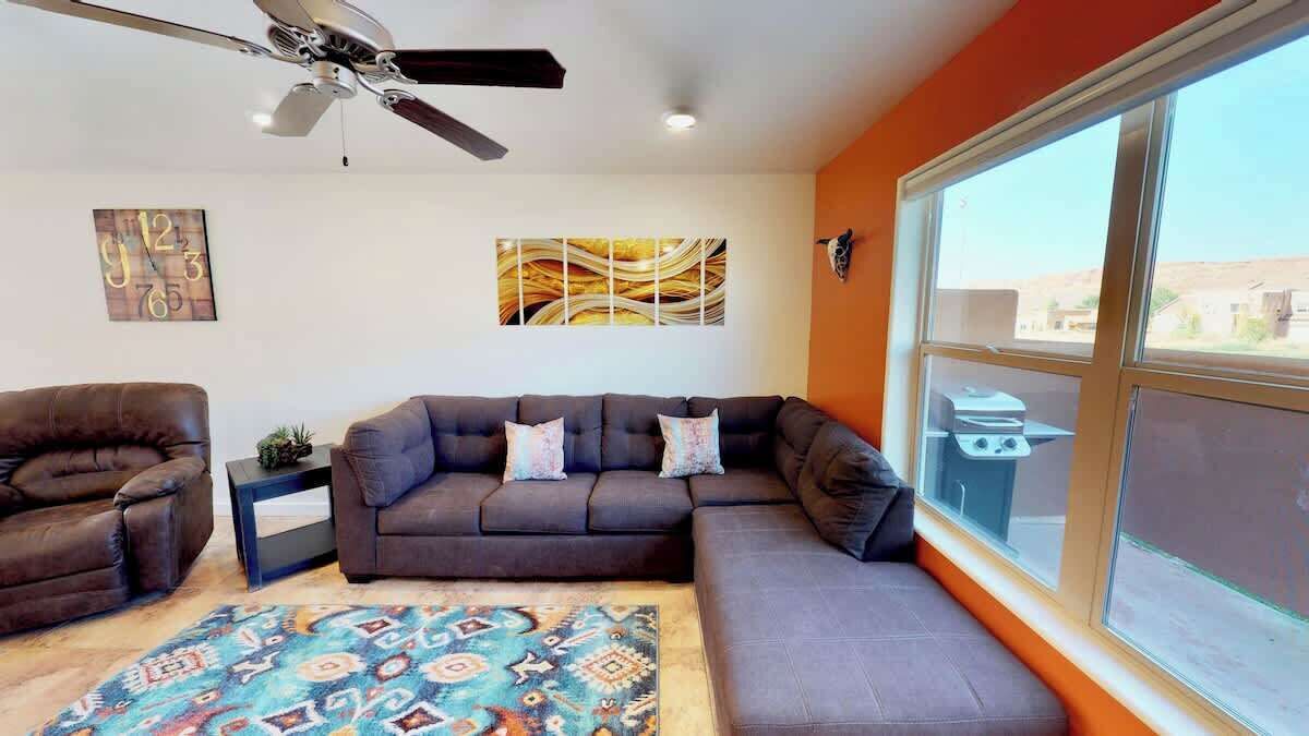 Una sala de estar con un sofá modular, un sillón de cuero marrón, un tapete estampado, un ventilador de techo y un ventanal con vistas.