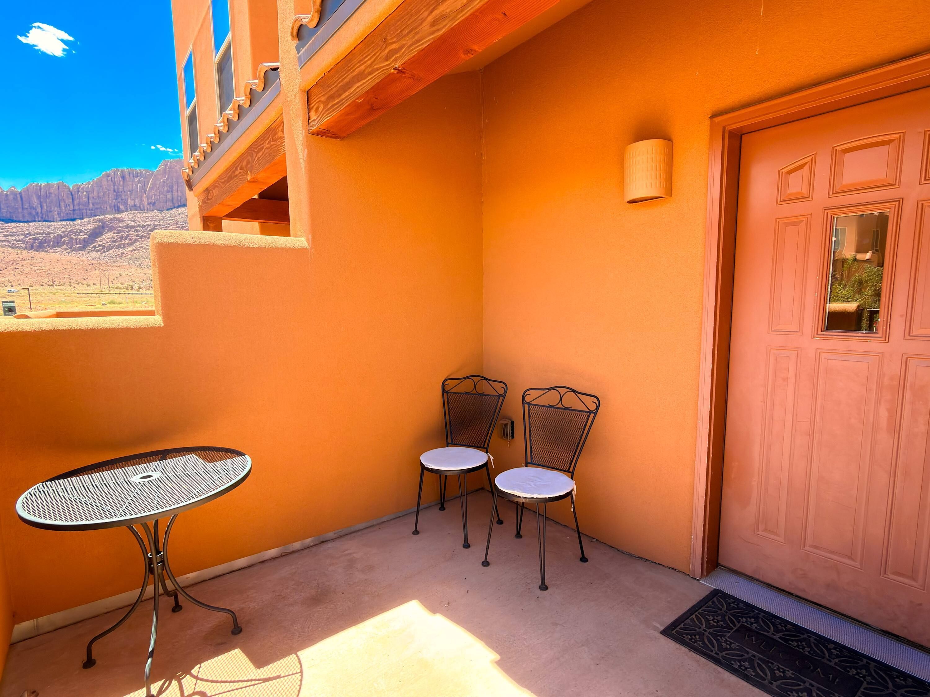 Un petit espace extérieur aménagé avec une table en métal et deux chaises, une porte en bois et une vue sur un paysage désertique.