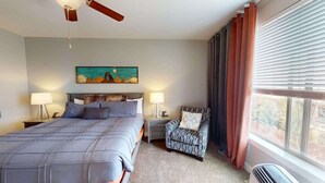 Un dormitorio con cama, dos mesitas de noche, una silla, ventilador de techo, ventana con cortinas y un cuadro en la pared.
