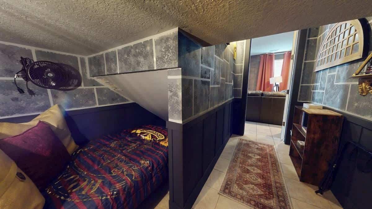 A room with a bed, a fan, a rug, and a bathroom in the background.