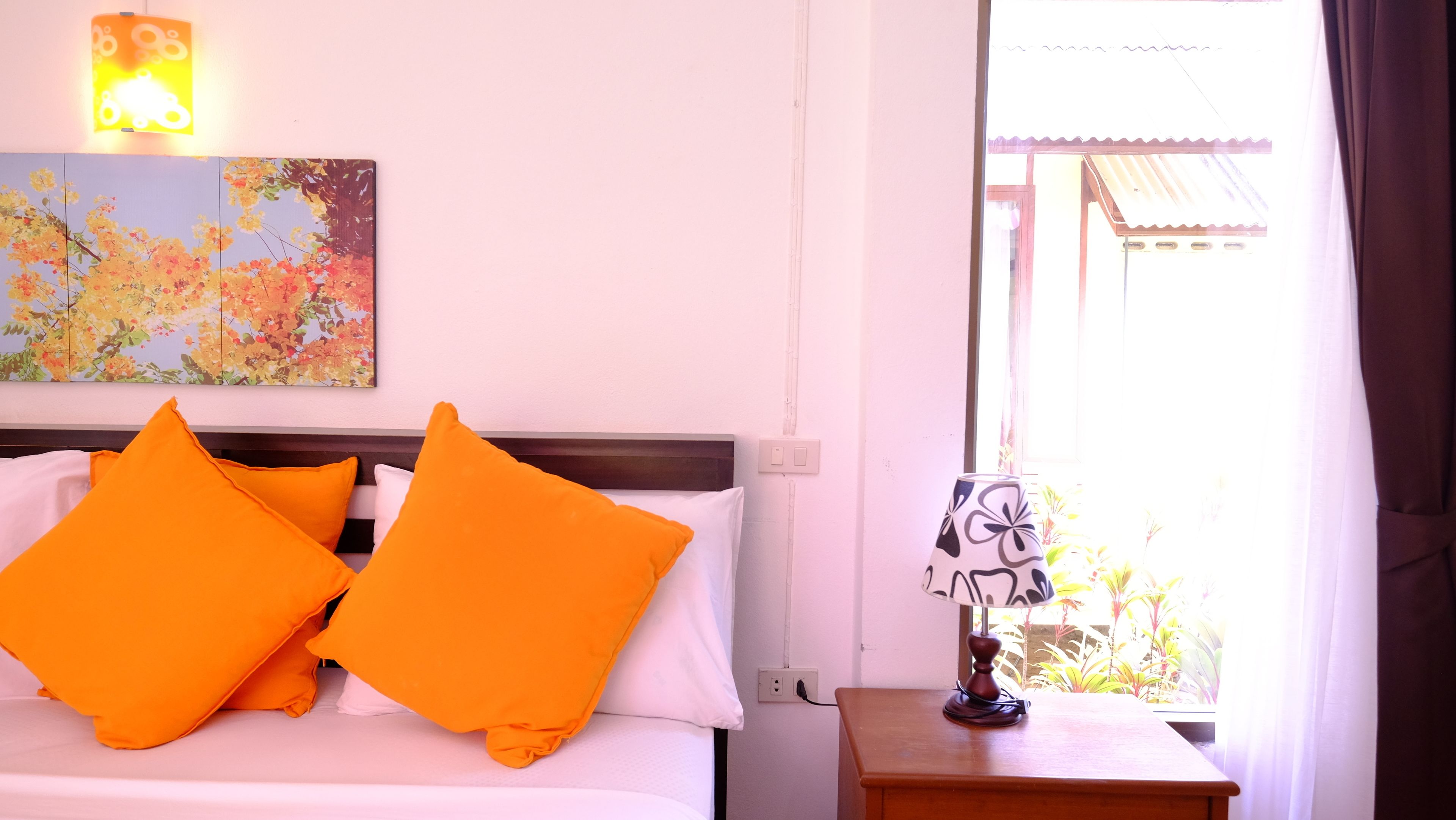 A hotel room with a bed, two orange pillows, a nightstand with a lamp, a wall-mounted light, and a window with a view of greenery and a building.