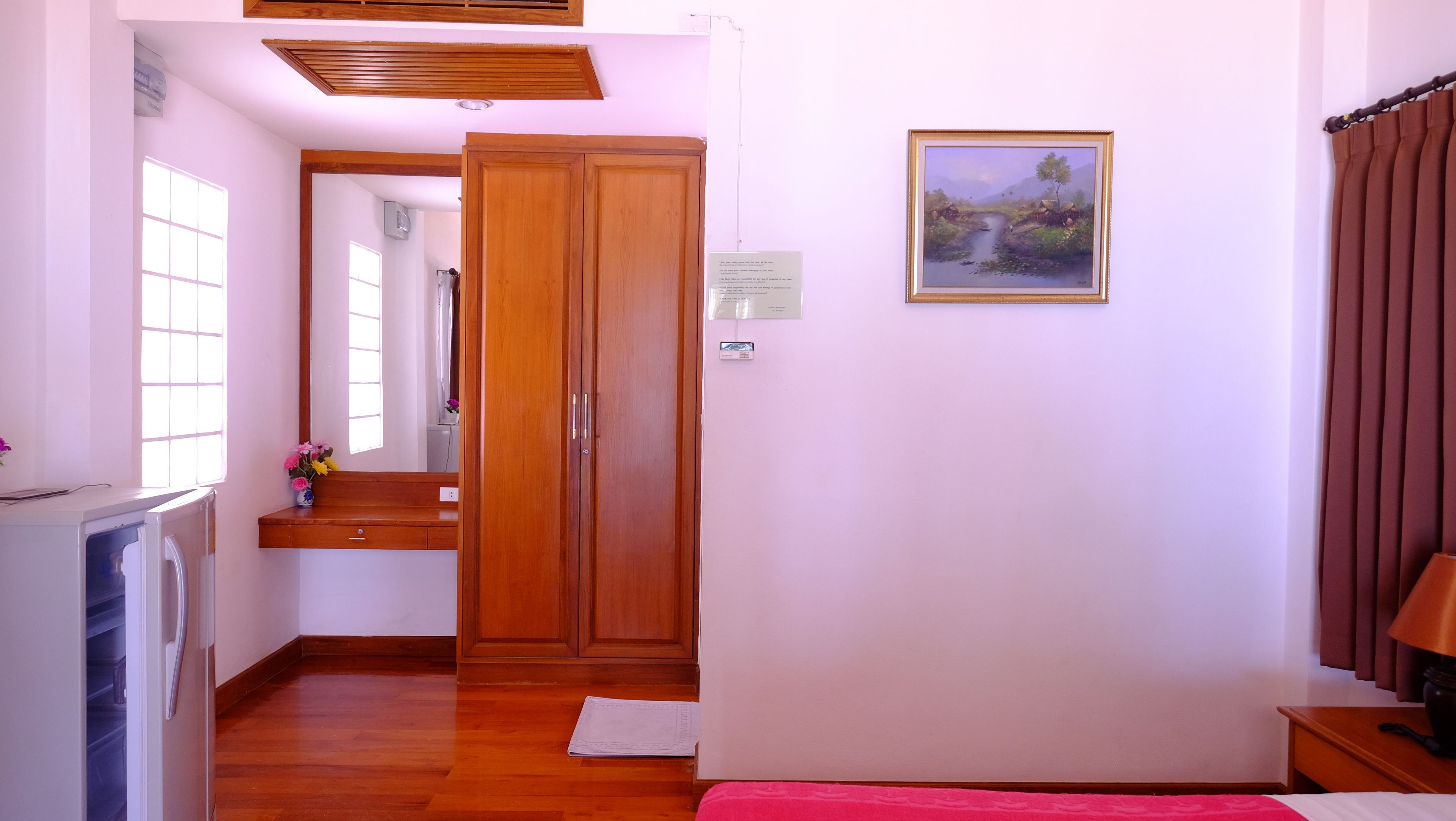 Une pièce avec une armoire en bois, un lit recouvert d’une couverture rose, une petite table avec une lampe et un tableau accroché au mur.