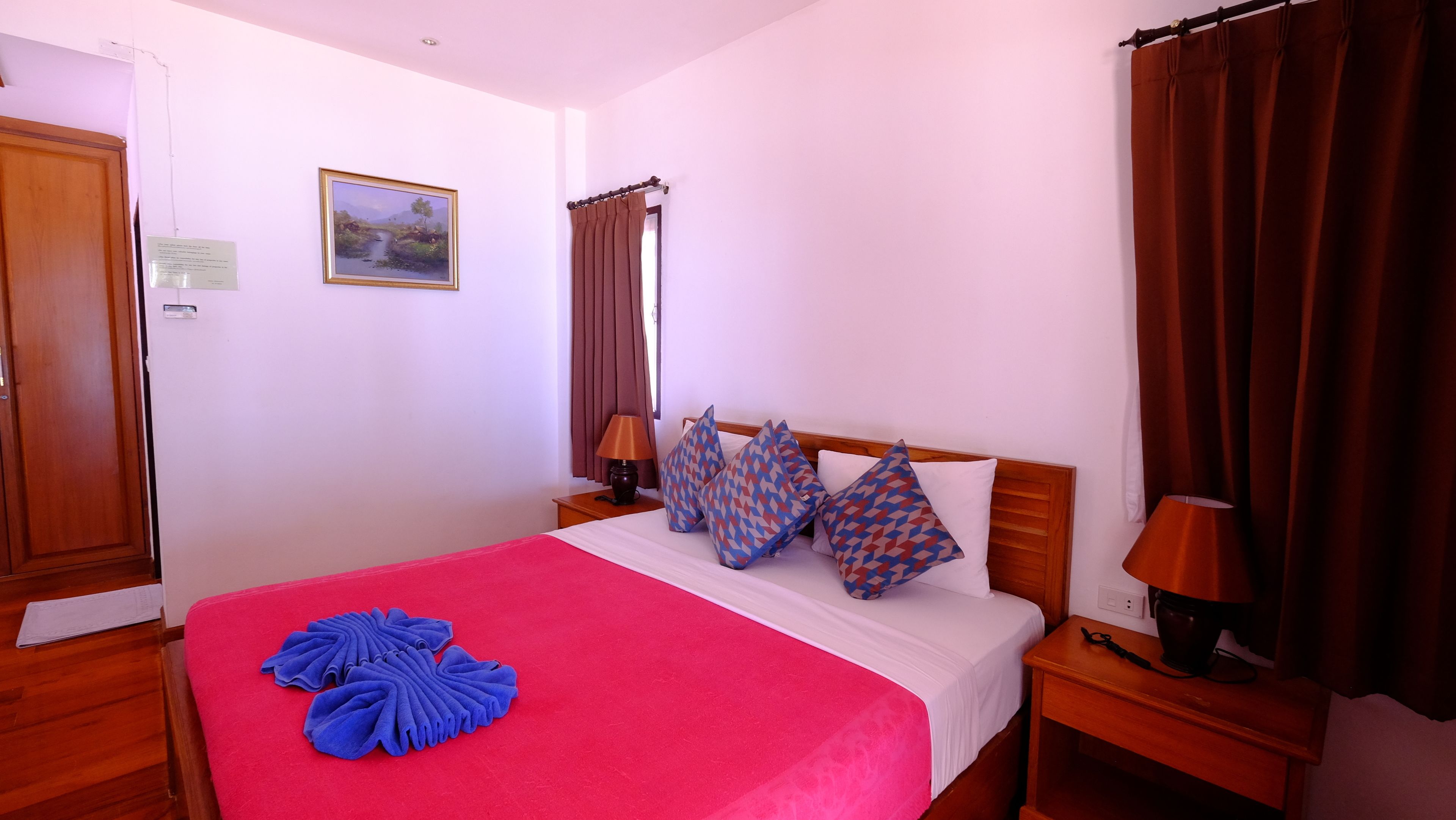 Une chambre d’hôtel avec un lit en bois, une couvre-lit rouge, des coussins à motifs, une table de chevet avec une lampe et un tableau accroché au mur.