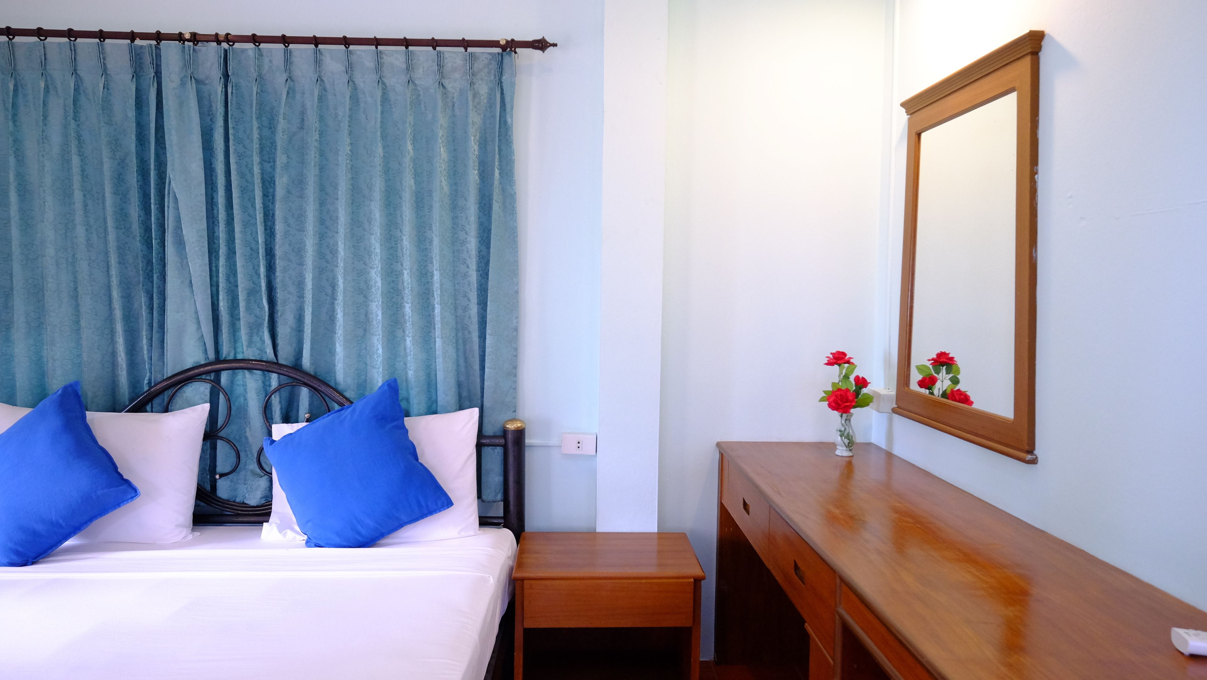 Un dormitorio con cama, mesita de noche de madera, espejo y ventana con cortinas azules.