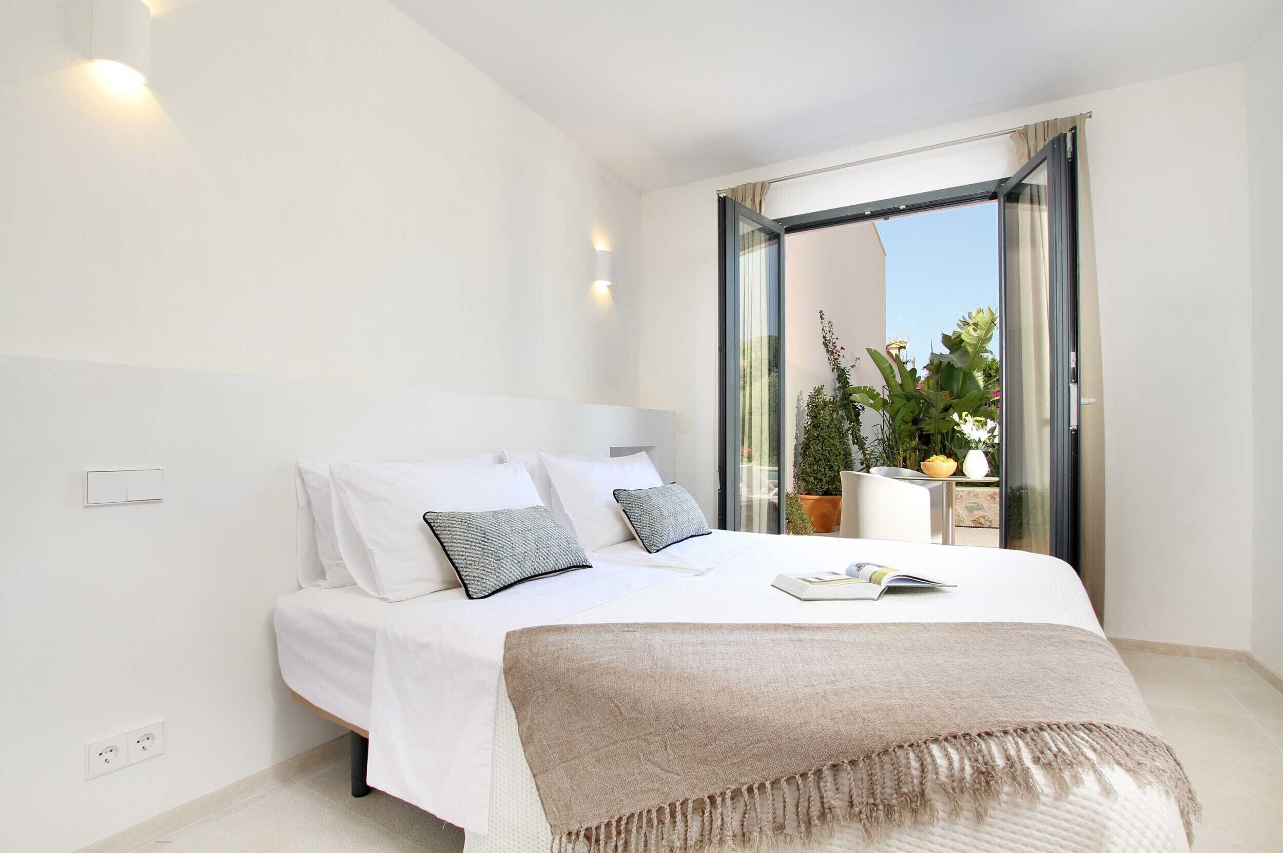 A modern bedroom with a large bed, white walls, and a sliding glass door leading to a balcony with plants.
