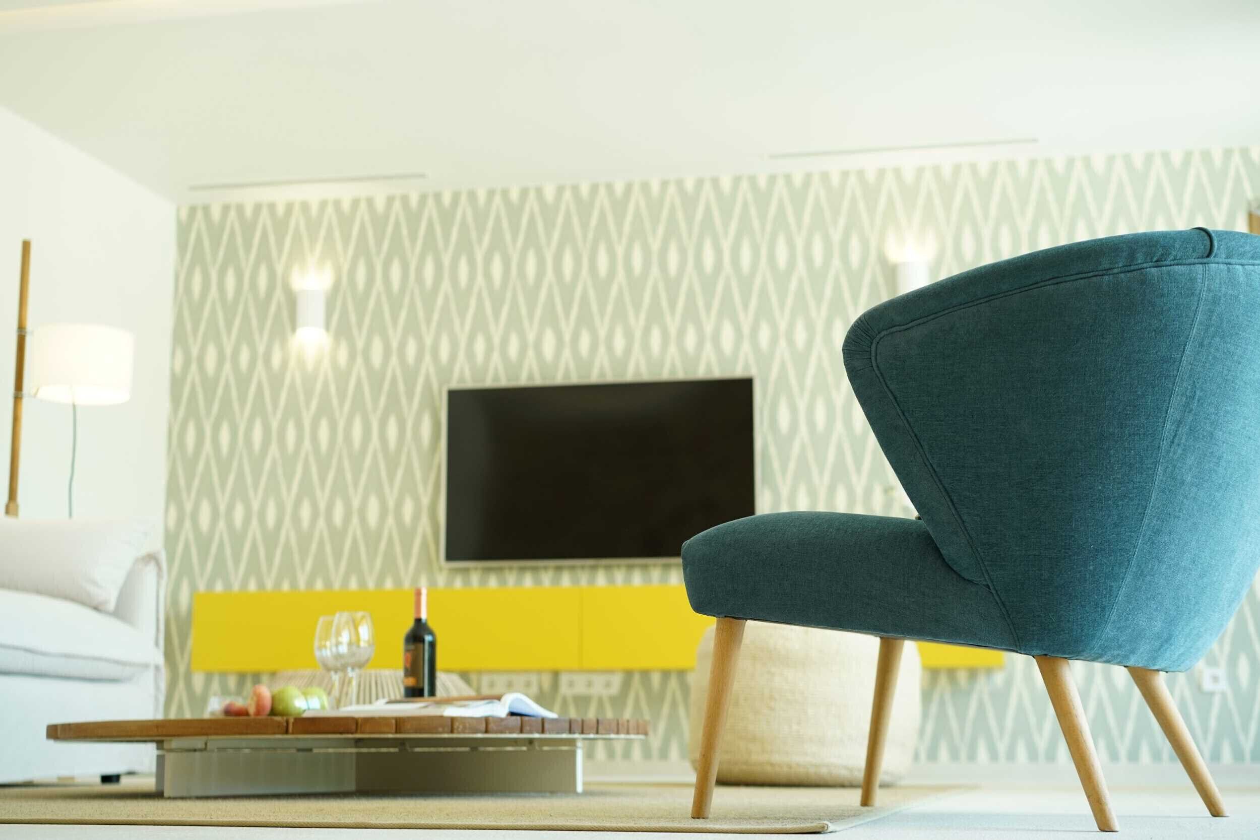 A modern living room with a teal armchair, a flat-screen TV mounted on a patterned wall, and a yellow accent wall.